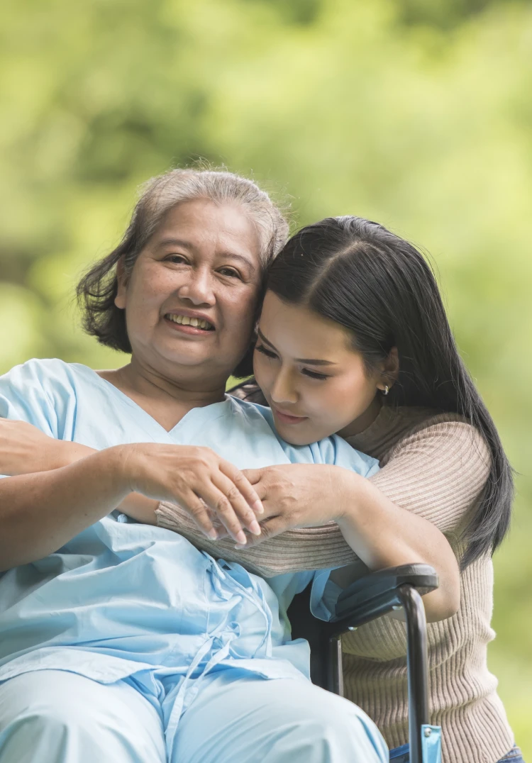 An elderly woman in a wheelchair embracing a younger woman, likely her daughter or granddaughter, in an outdoor setting surrounded by greenery.